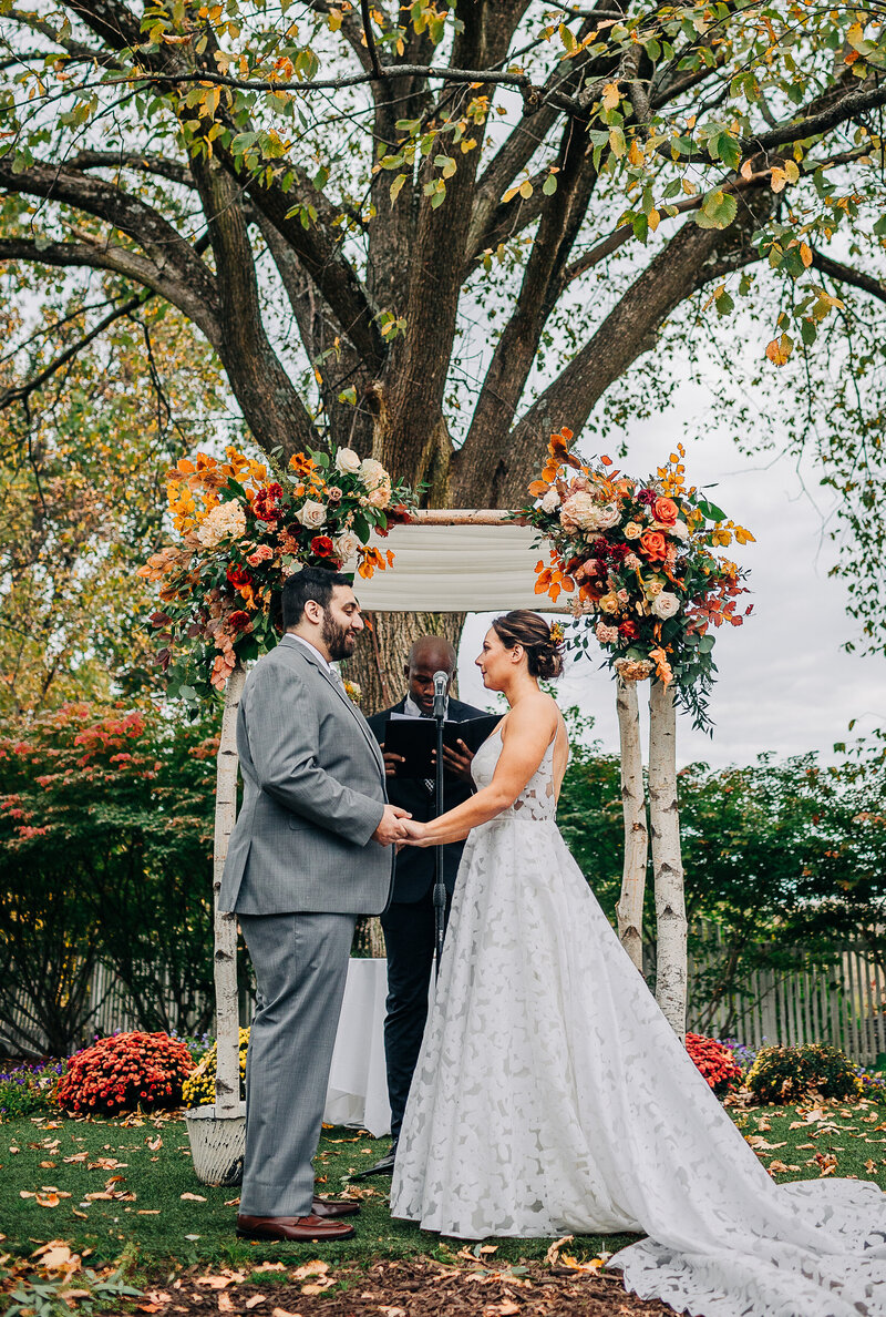 A bride a groom and their officiant exchange vows in New York's Hudson Valley before a birch chuppah decorated with large autumn flower arrangements