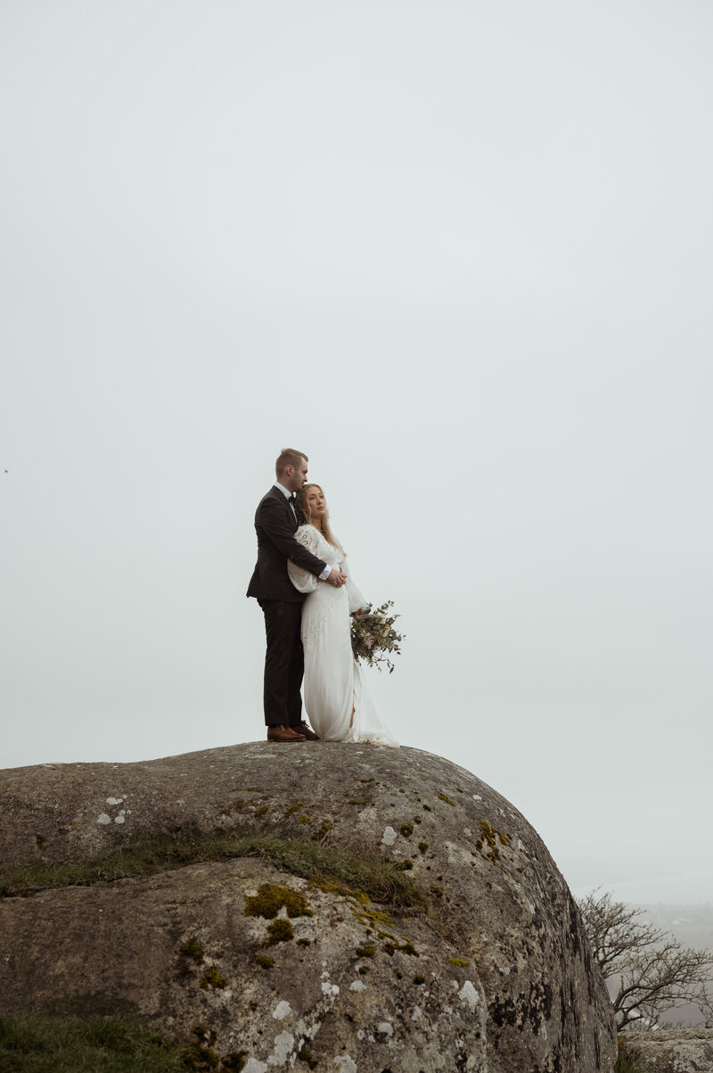 Brudpar står i regnet och kramas på en klippa