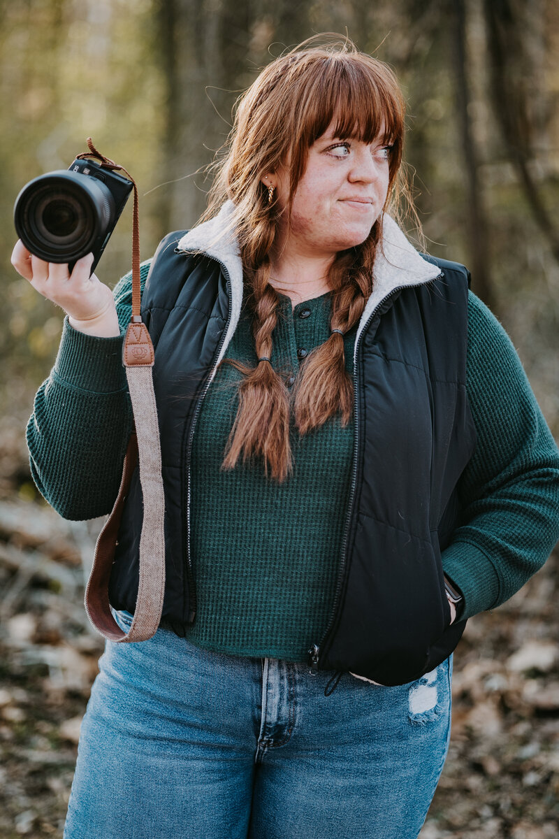 Savannah holding her camera and smiling off to the side, with the sun shining through the trees behind her, creating a warm, natural glow.