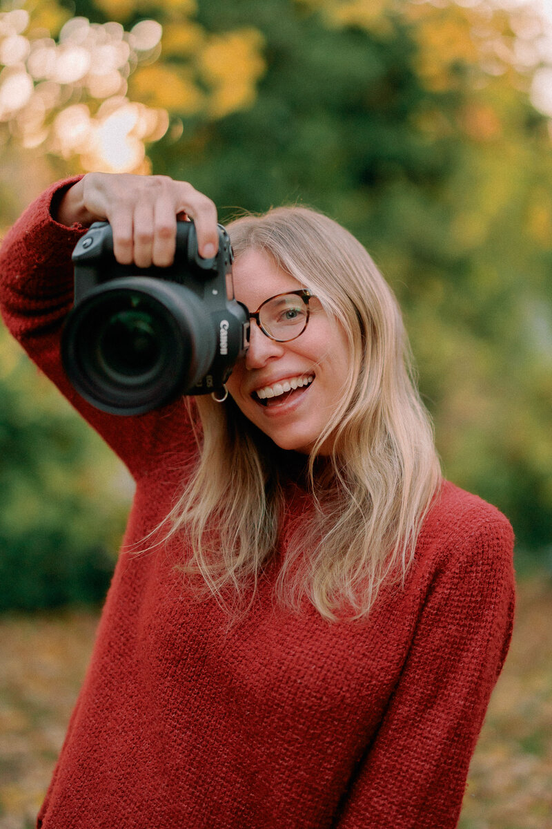 A photographer smiling while holding a camera up in one hand 