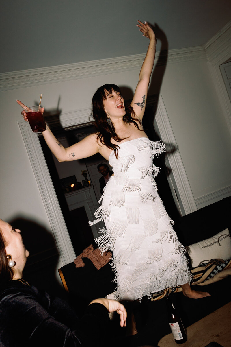 Bride standing on couch in tassle dress with drink in her hand having fun at her wedding in Portland, Maine