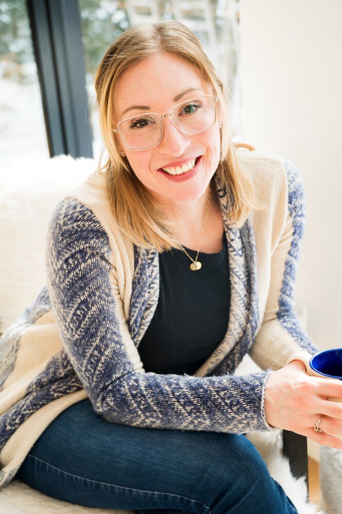 A woman with glasses holds a blue mug while sitting on a couch, representing an anxiety therapist in Ontario.