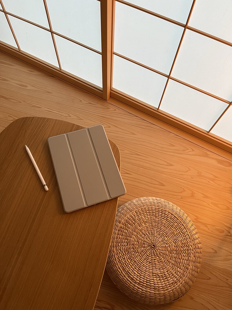 A serene setting with a wooden table holding a tablet and stylus, next to a woven cushion. Shoji screen filters warm light, creating a cozy atmosphere.