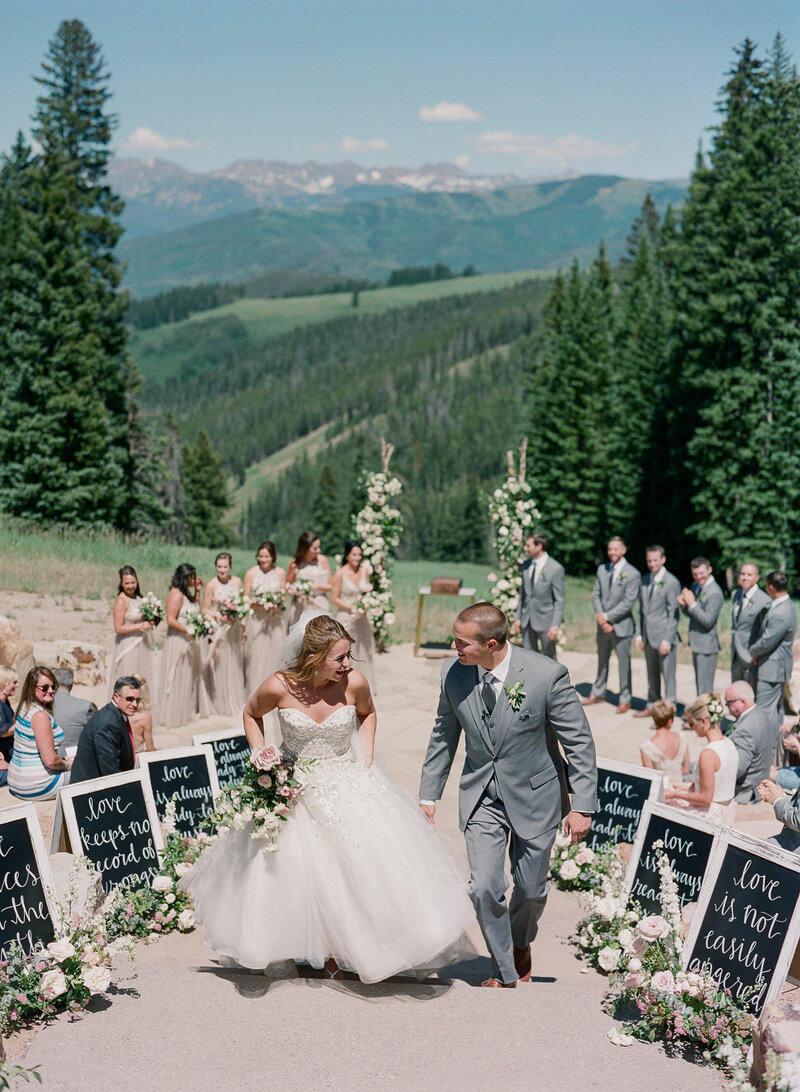 Empty outdoor wedding ceremony space, lined with wooden chairs. There is a white aisle, with a floral chuppah at the end, there are mountains in the background.