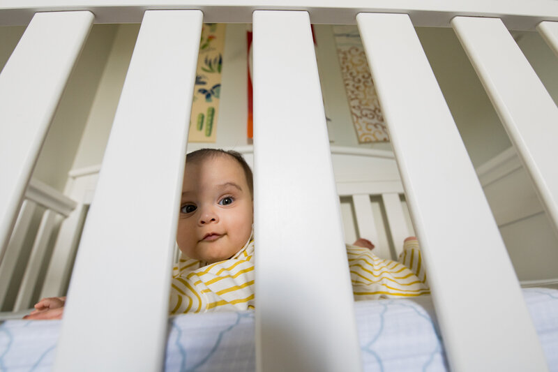 a newborn baby looks through the white bars of his crib wearing a yellow onesie