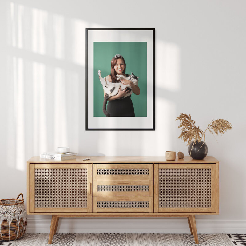 A picture of a wall in a home. Includes a picture in a frame of a woman wearing a tiara and holding a cat.