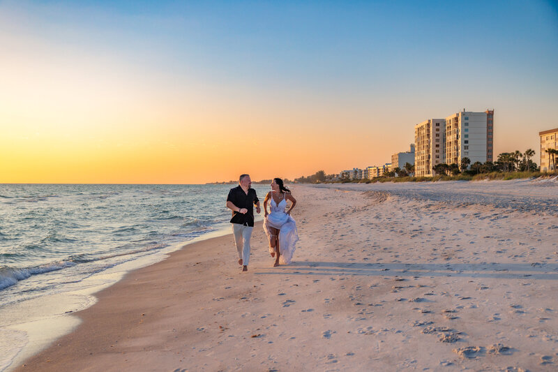 Bride and groom running towards the camera on the beach looking at each other with the sunset behind them.