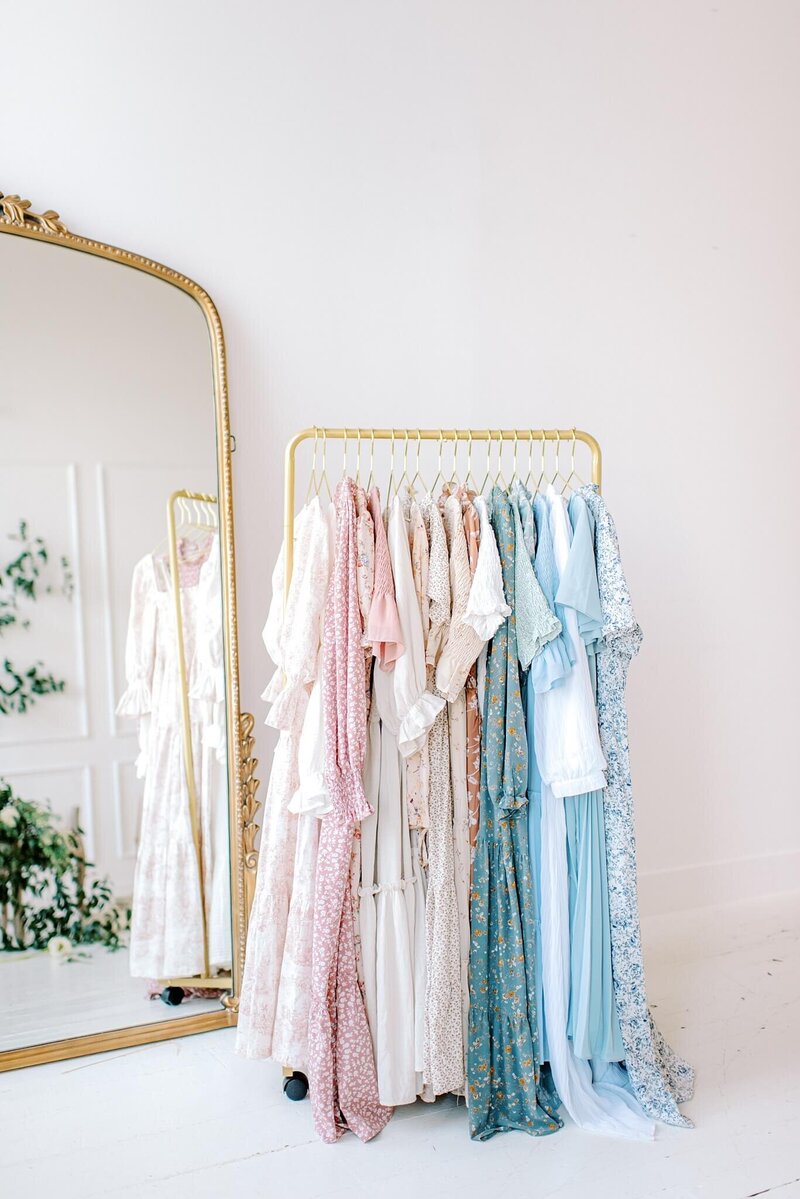 Dresses from Melissa Mayrie Photography's client closet hang on a gold wardrobe rack placed in front of a large Anthropologie mirror