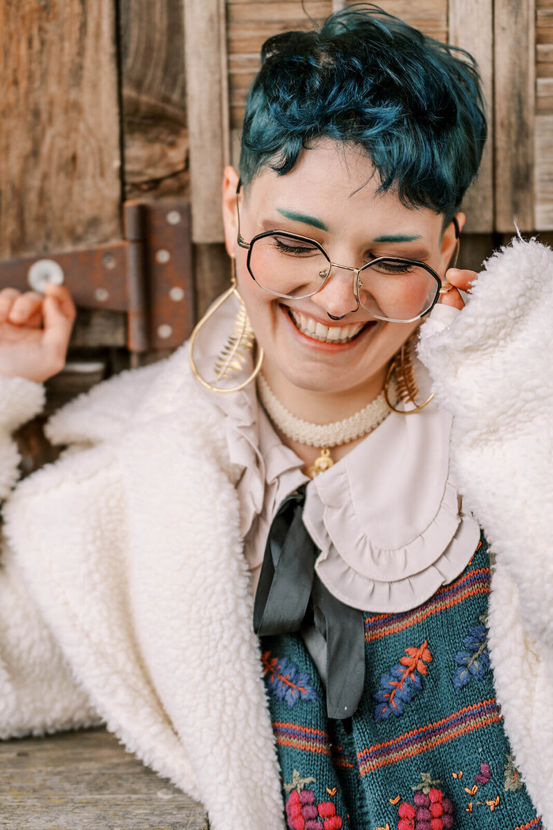 A person wearing glasses smiles  authentically, representing the shooting style of lifestyle photographer Among the Oaks.