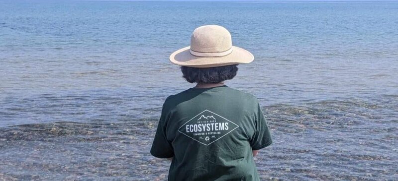 A woman wearing a hat is seen from behind wearing an Ecosystems shirt looking at water, representing the company's commitment to environmental stewardship.