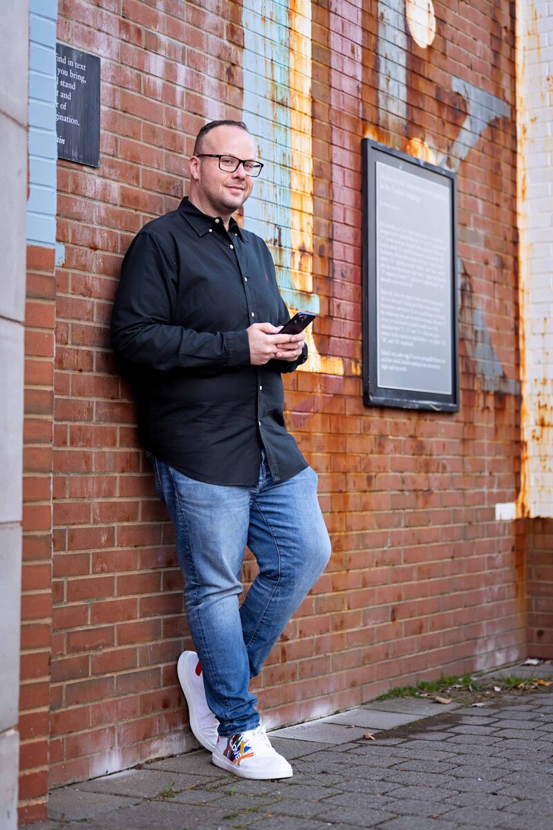 Dr. Mike Stokes standing outdoors against a brick wall with a phone in hand, smiling confidently.