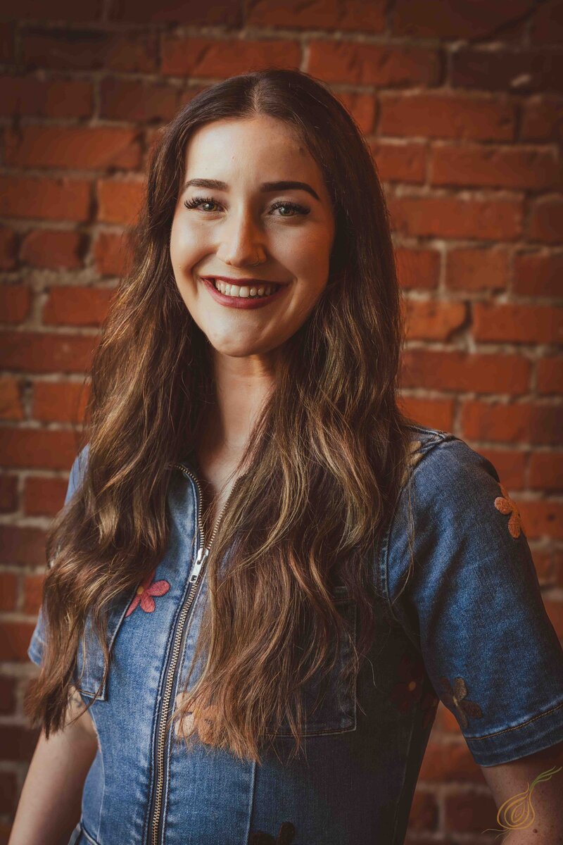 A cheerful woman stands against a brick wall, her smile conveying warmth and happiness.