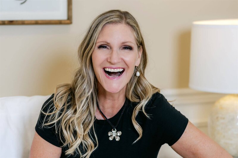 Phillipa Leseberg with long blonde hair smiles broadly, wearing a black shirt and a flower-shaped pendant necklace. A lamp is visible in the background, hinting at the cozy setting where author Phillipa Leseberg crafts her memoirs.