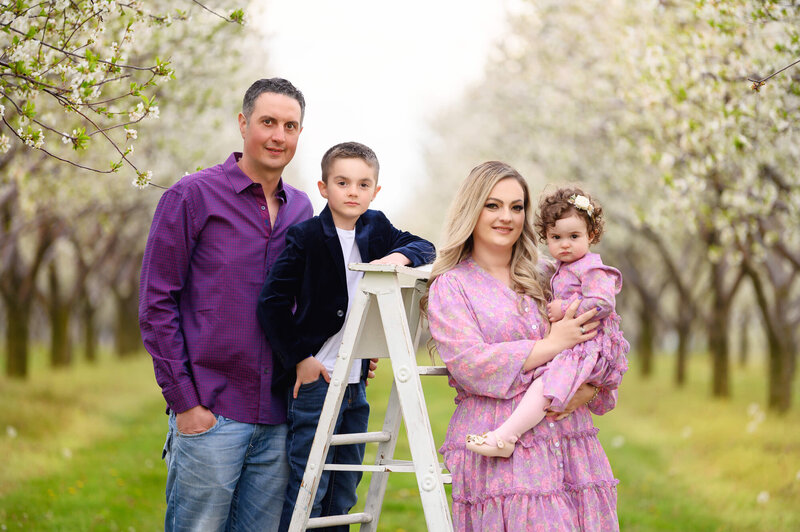 Mom and dad standing with their son and daughter leaning against a white ladder in a cherry blossom field for their Spring family photos in Burlington, On