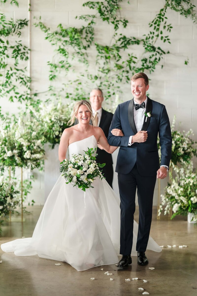 A bride and groom beam as they walk down the aisle surrounded by green florals