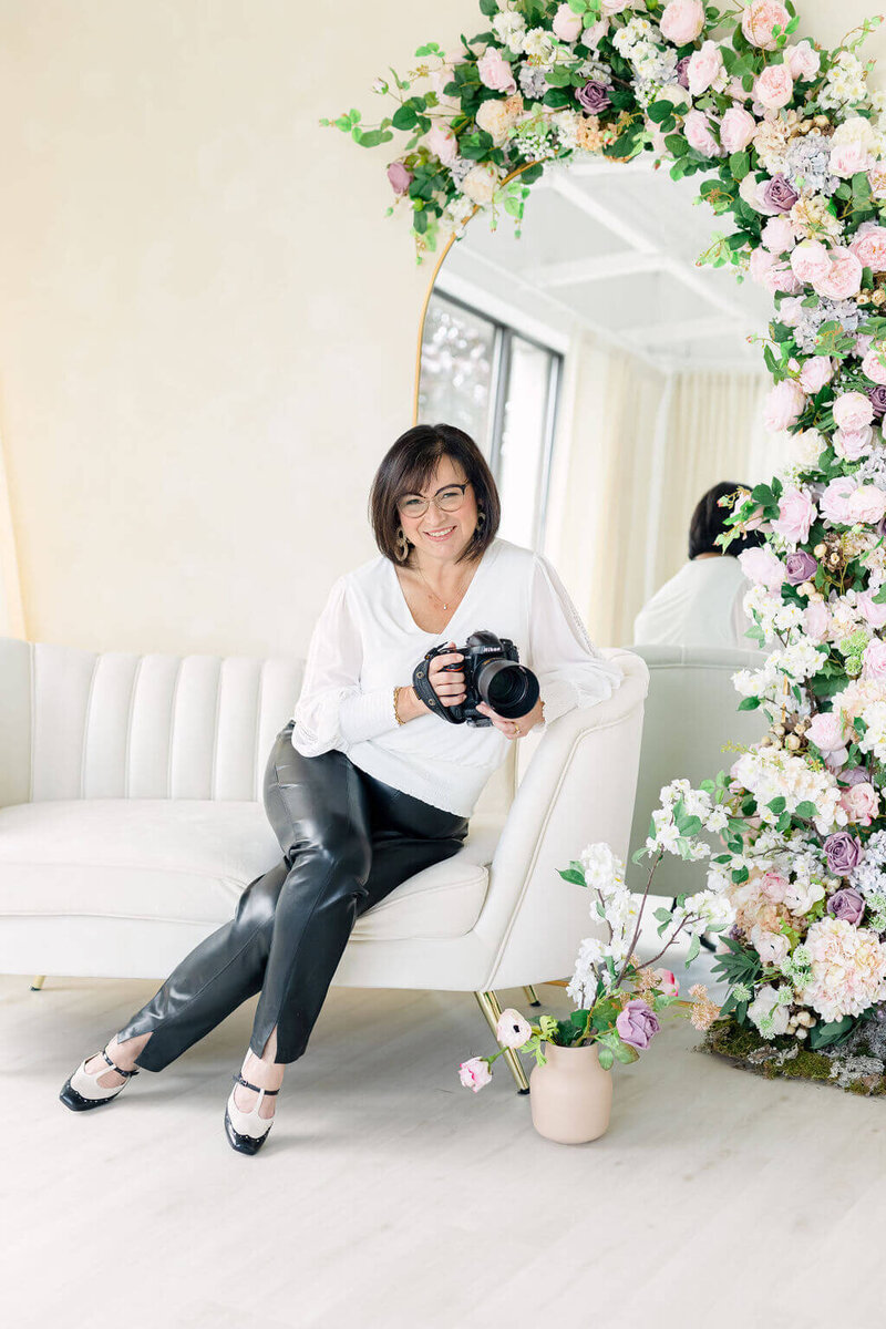 photo of Ottawa Wedding Photographer Elenora Luberto JEMMAN Photography sitting on a couch holding a camera with a mirror and floral arch behind her