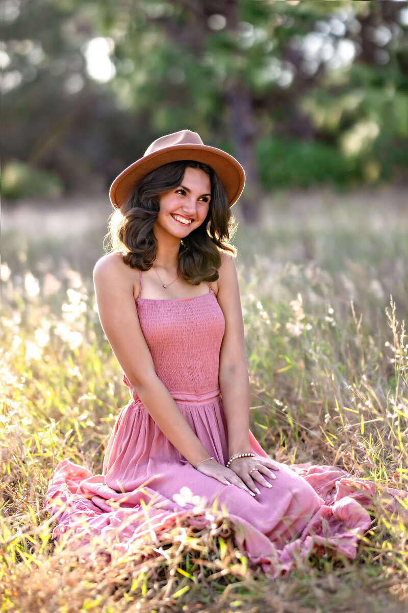 Senior girl in pink dress and cute hat smiling softly while looking away in gorgeous golden hour sunlight in Winter Garden Florida