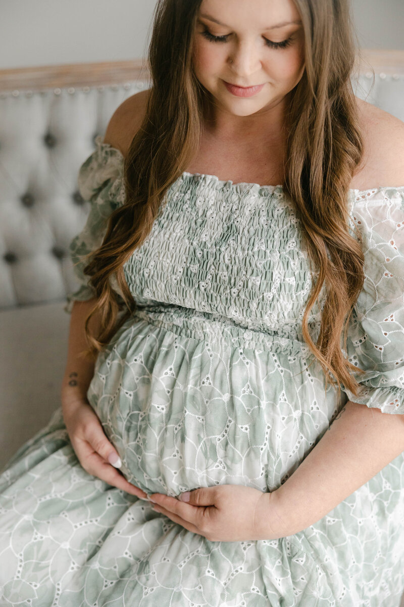 mother holds her pregnant belly while sitting in a sage green floral dress, Indianapolis  maternity photographer
