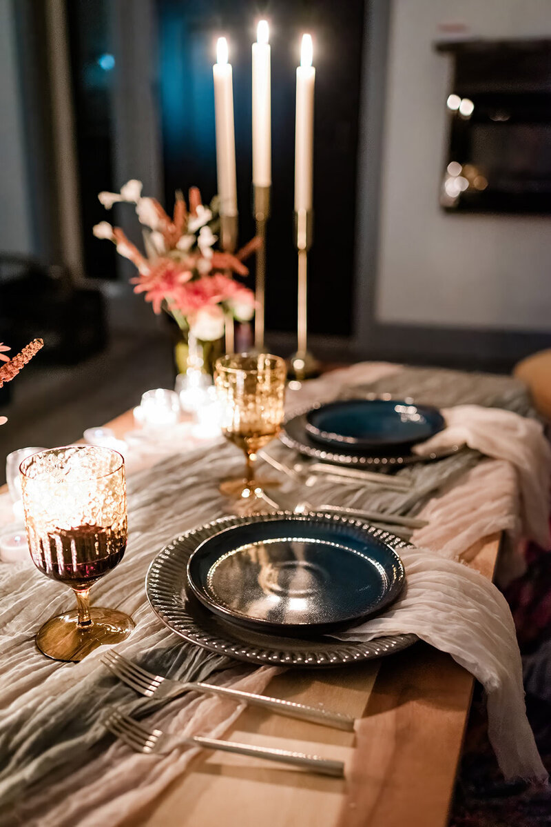 A beautiful, simple sweetheart table styling at a a private chef prepared dinner for an eloping couple as part of their Denali Glacier Landing elopement package.  There are flowers in on the table, soft green and cream colored linens set next to the blue and green earthy looking plates.  The table for two is lit in a warm candle light glow as there are many candles all across the table. 
