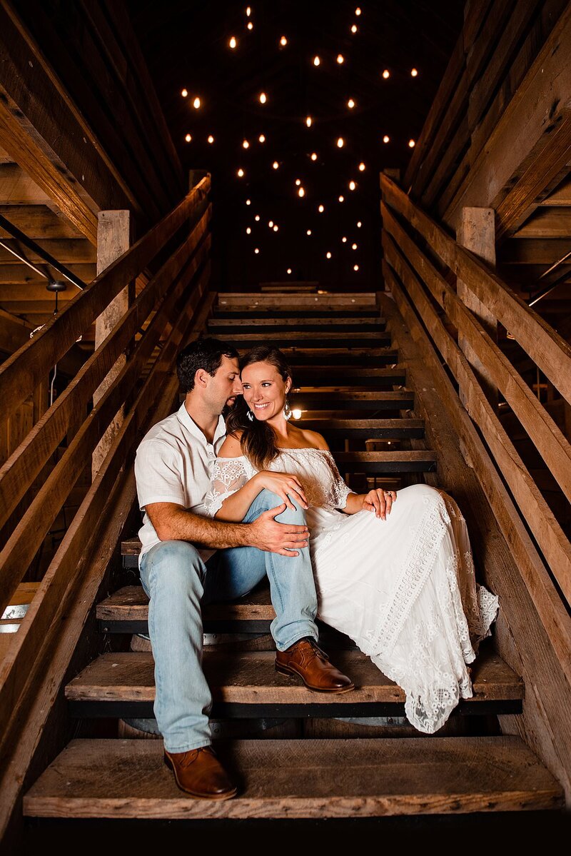 The bride and groom sit at the bottom of a wooden staircase. The groom, wearing brown shoes, light blue jeans and a white short sleeved golf shirt, whispers into the bride's ear. The bride leans against him and laughs at what he is whispering to her.  The bride is wearing an off the shoulder lace boho maxi dress with a wide lace ruffle around her shoulders. The wooden railing for the stairs is on either side of them and the string lighting at the top of the stairs looks like stars in the night sky.