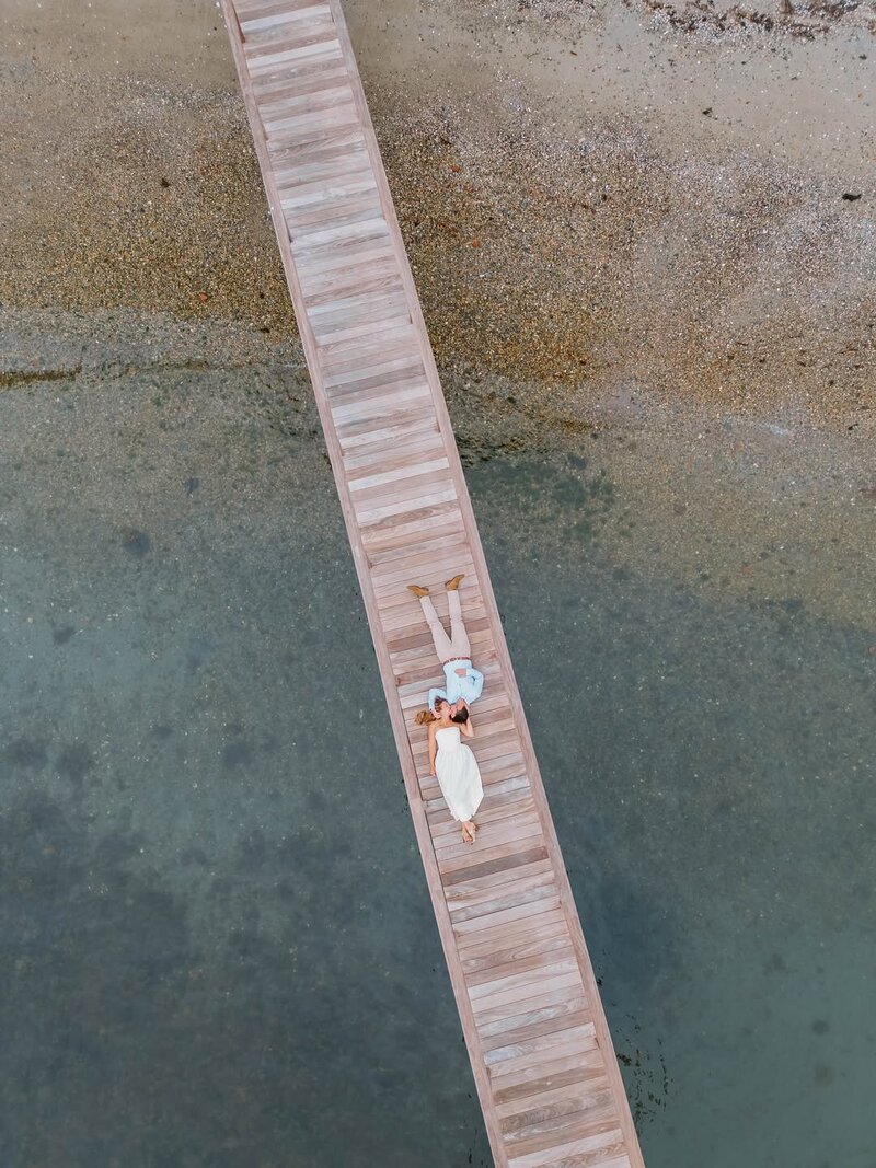 Rhode Island Wedding photographer | Aerial view of a couple lying on a wooden pier above clear, shallow water and pebbly shore. 