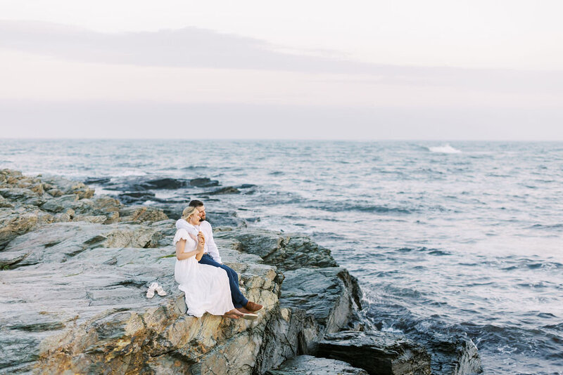 Bride and groom hold each other sitting on large rocks by the ocean in Newport, Rhode Island