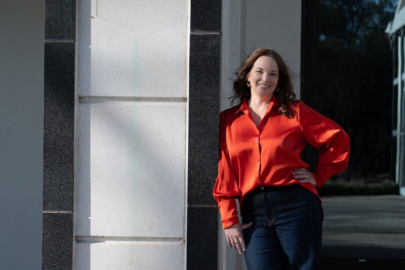 brown haired woman smiling, wearing a red shirt and leaning against a building outside