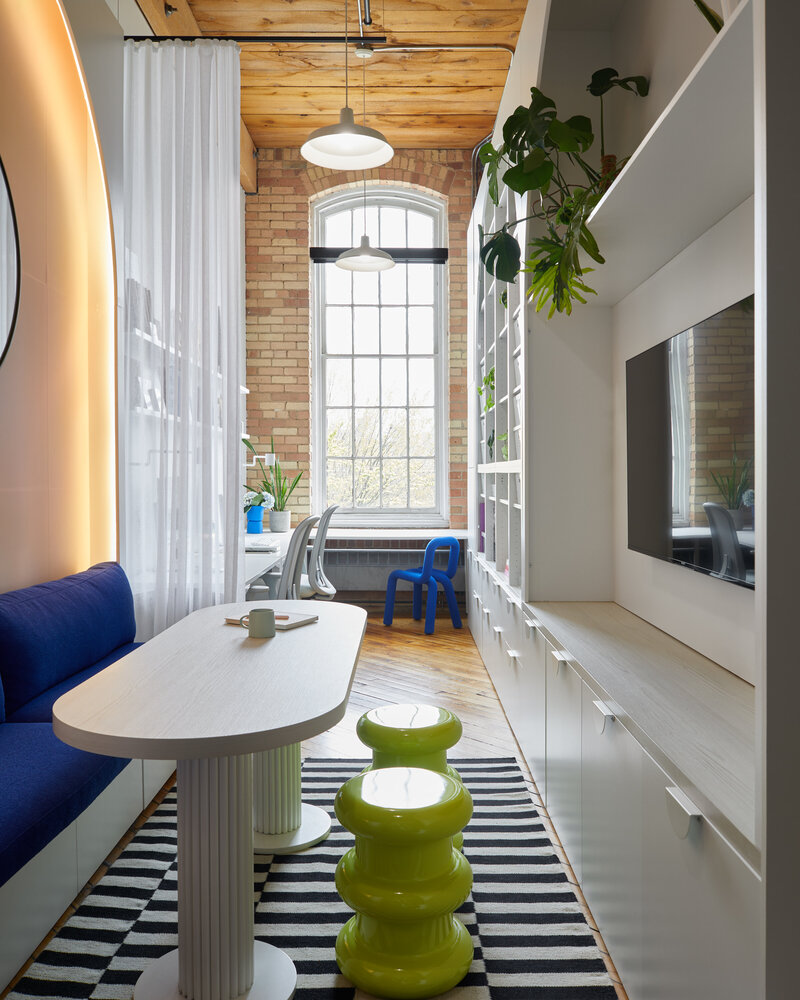 A tall, narrow office space containing an elegant wood table surrounded by stylish blue and green chairs on top of a black and white rug with a wall-mounted TV screen on the opposite wall. A sheer white curtain partition partially hides a modern office space containing two white chairs and desks, potted plants, a tall paned window, and large white shelves