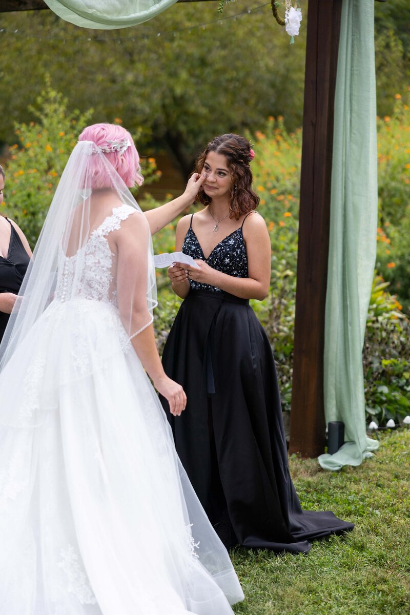 Pink haired bride wearing lace and tool wedding dress wipes away tear on bride wearing black beaded wedding dress