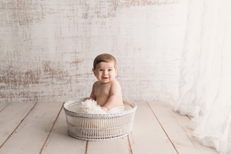 An infant baby sits and plays in a white wicker basket on the floor of a studio run by a Lafayette Baby Milestone Photographer