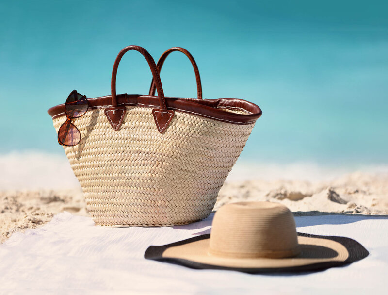 Straw beach bag with sunglasses and a sun hat resting on white sand by the ocean.
