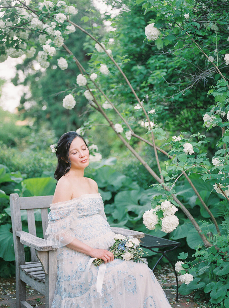 Asian mother with blue Needle & Thread dress sits on outdoor chair in garden next to hydrangea bush.