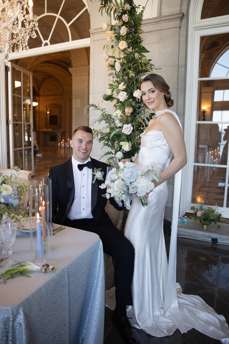 Aldrich Mansion Wedding Photos | Kelsey Sheehan Photography Timeless Rhode Island Weddings | A bride in a satin gown stands beside a seated groom in a tuxedo. They smile near an elegant table, with lush floral arrangements and soft lighting.