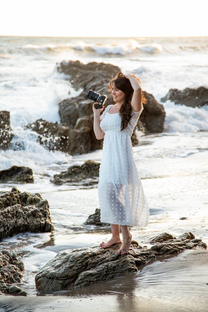 Heidi holding camera with one hand and touching hair with other standing on rock on California beach | Maine Wedding and Portrait Photographer