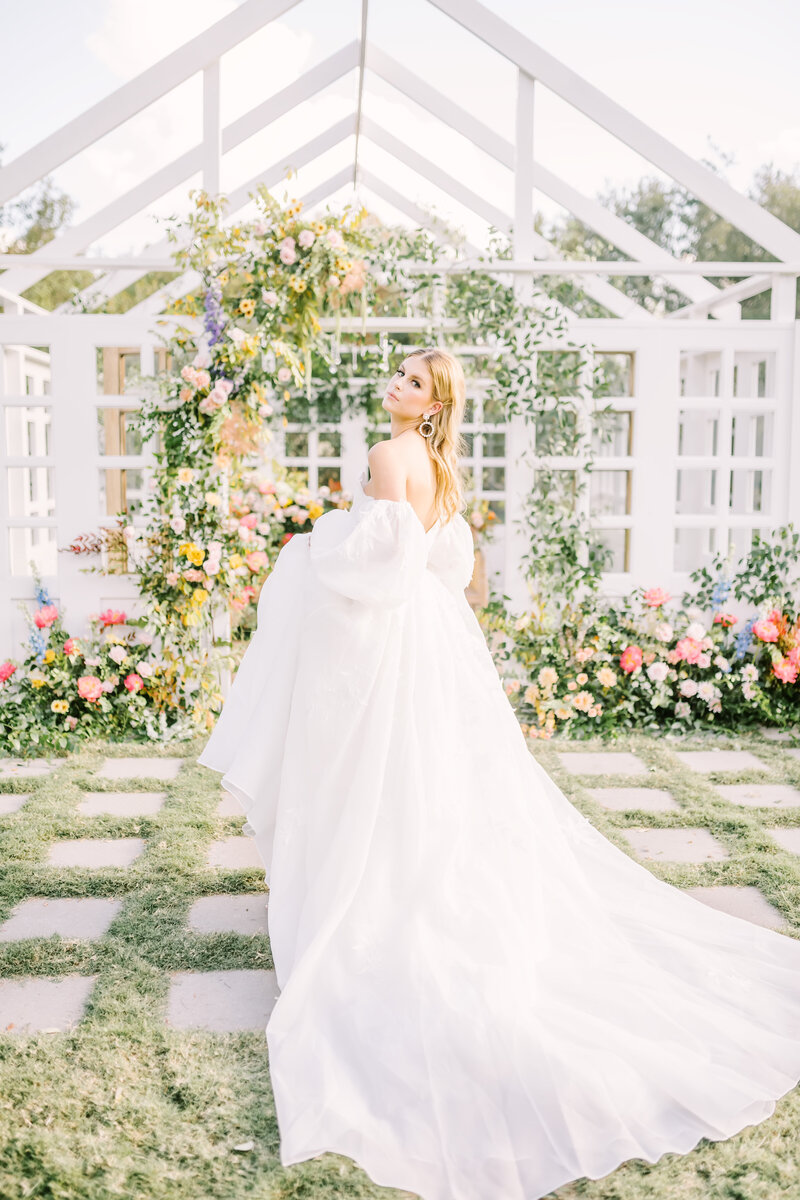 Gorgeous bride looks over her shoulder in her wedding gown in front of trellis decorated with lush florals