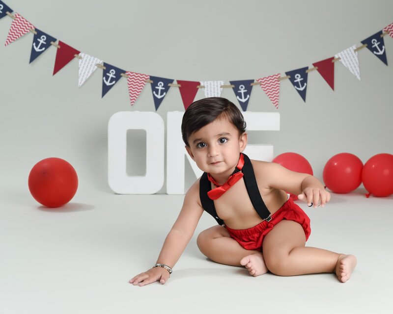 A baby boy in a red and black outfit with a bow tie crawling on the floor, surrounded by red balloons and nautical-themed decorations that spell "ONE."
