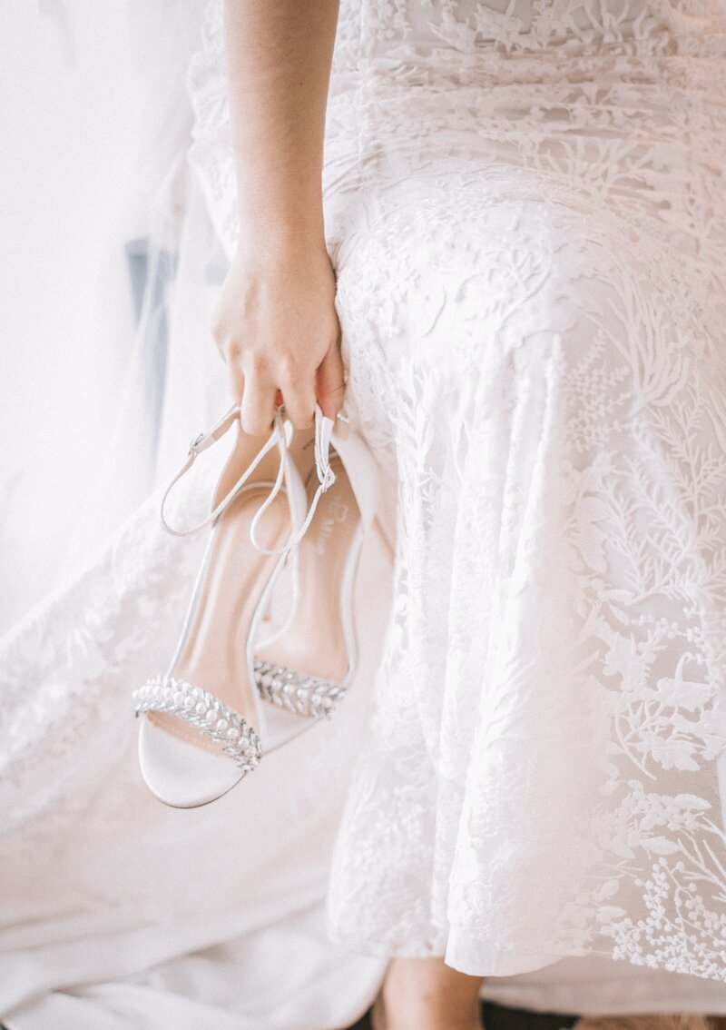 A bride in a lace gown holding her shoes adorned with pearls and crystals.