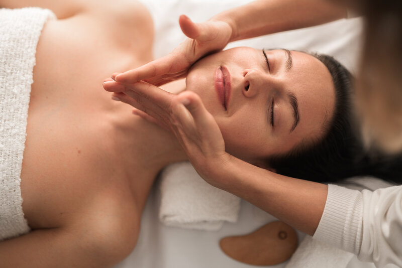 Gentle facial massage on a relaxed woman with gua sha tool visible, promoting facial sculpting and relaxation.