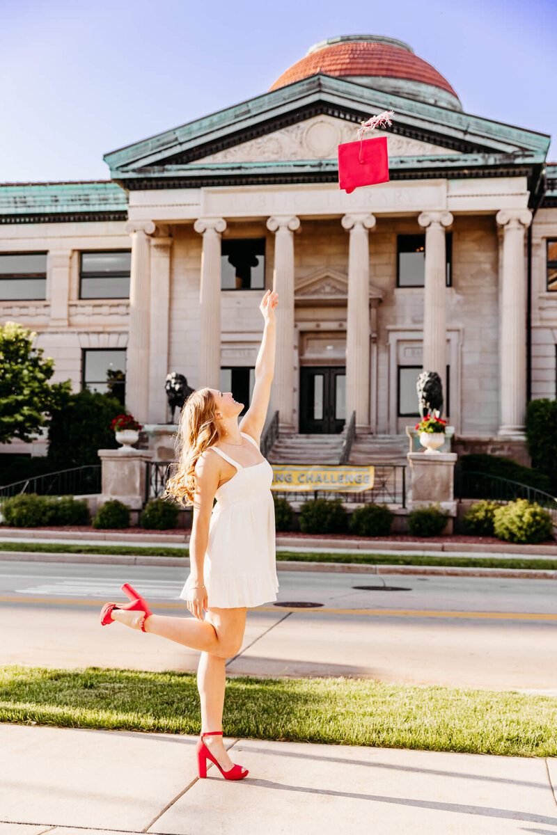 Beautiful brown haired woman crossing her ankles and touching her elbow while wearing her cap and gown for her graduation photo session in Green Bay