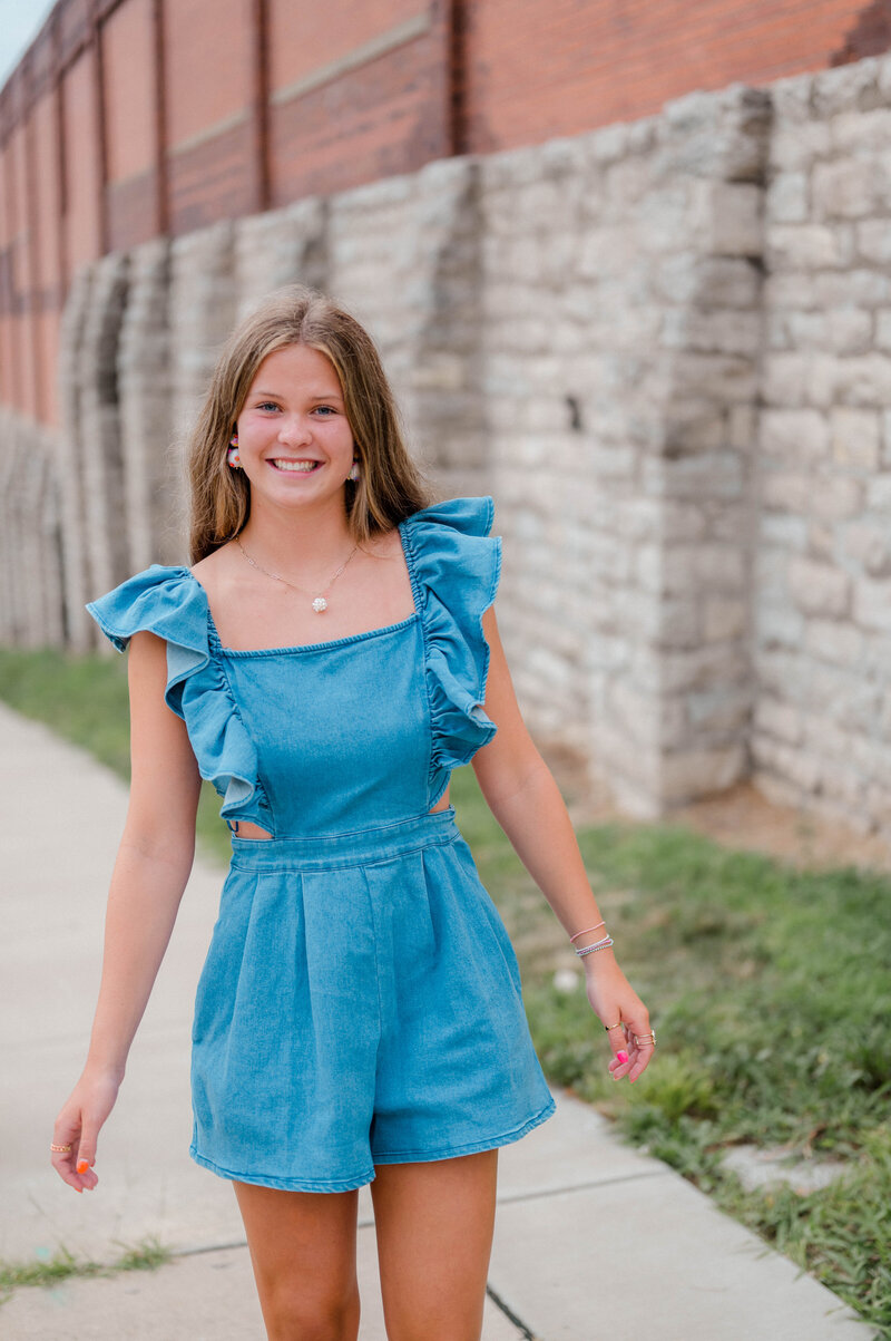 High school senior wears blue romper and walks forward smiling at the camera.