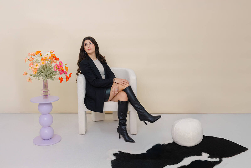 Bel, the founder and owner of Soulve Studio, sits elegantly in a white chair wearing a black blazer, leather skirt, and knee-high black boots. She is posed thoughtfully next to a vibrant floral arrangement on a purple side table, creating a stylish and modern atmosphere against a neutral beige background. The scene reflects her creative and fashionable flair.