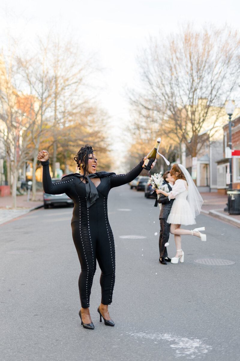 Black drag queen officiant wooing popping a champagne bottle with the nonbinary wedding couple seal their vows with a kiss in the background in the middle of the street in Georgetown DC 