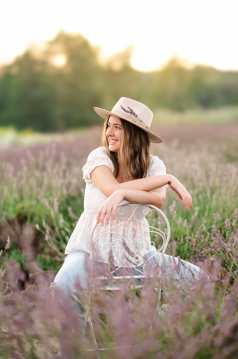 Senior girl in a soft dress and wide-brimmed hat sitting in a tall grassy field at sunset in Lancaster, PA