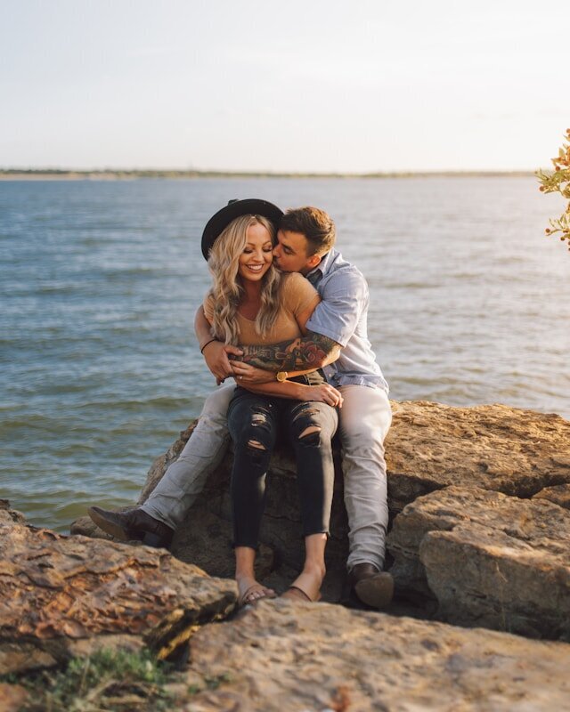A couple hugs while sitting on rocks next to water representing event makeup by Libby.