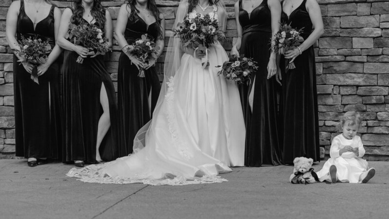 Grumpy flower girl sitting at the feet of the bridesmaids during portraits, completely unaware of the camera, adding a touch of humor.