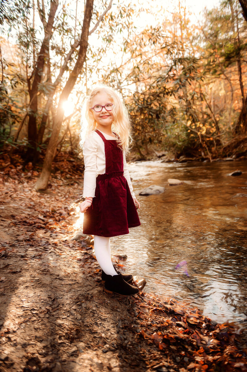 Little girls smiles by a creek in autumn during her family photoshoot in Asheville, NC.