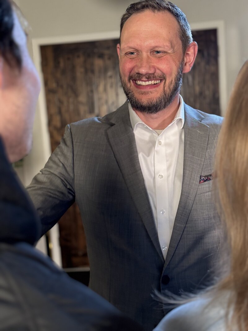 Man in a light gray suit with pocket square and no tie, smiling and talking to people