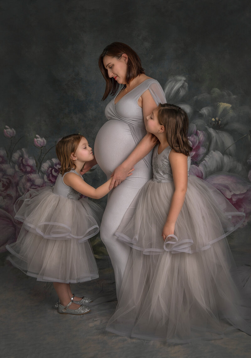 "Two daughters with their expecting mother in a studio maternity session, one kissing her mom's belly while the other looks at her sister in Ottawa"
