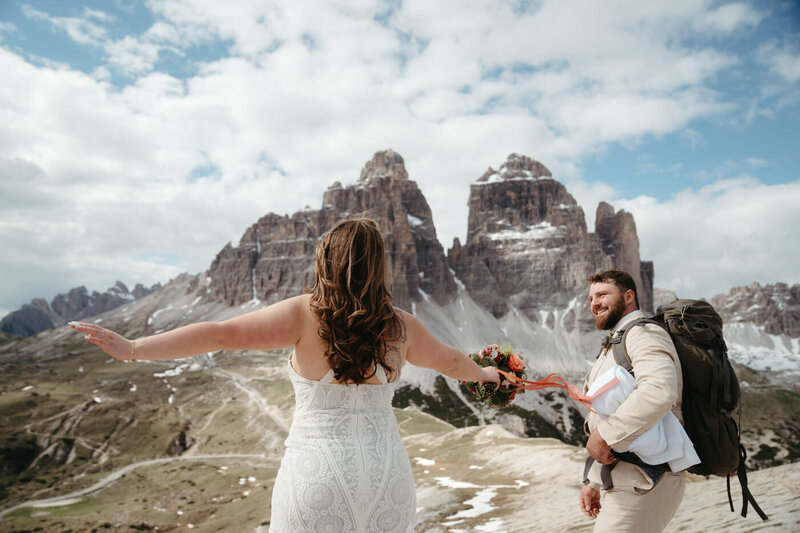 Dolomites Elopement | Groom smiles at bride as she dances with joy surrounded by mountains