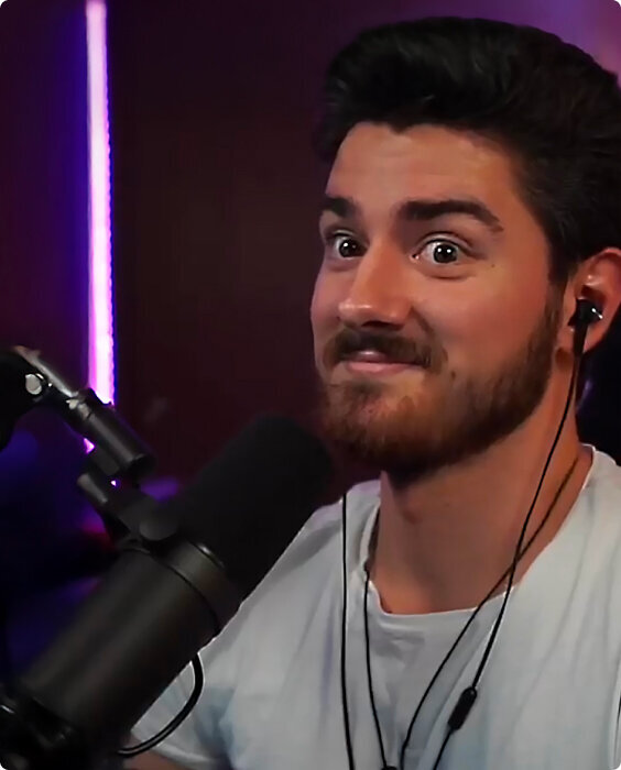 Man sitting in a purple-lit room with short brown hair and full beard and mustache. He is smiling slightly and wearing a white shirt. A microphone is propped next to his mouth.